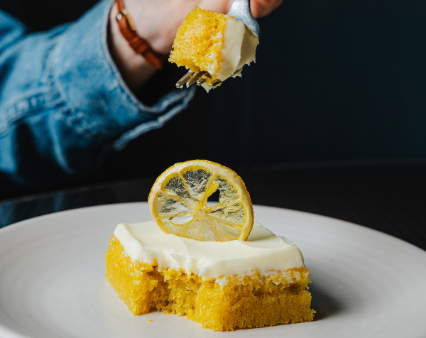 A fork with a bite of lemon sponge cake over a slice of cake with creamy white chocolate frosting and a candied lemon slice.